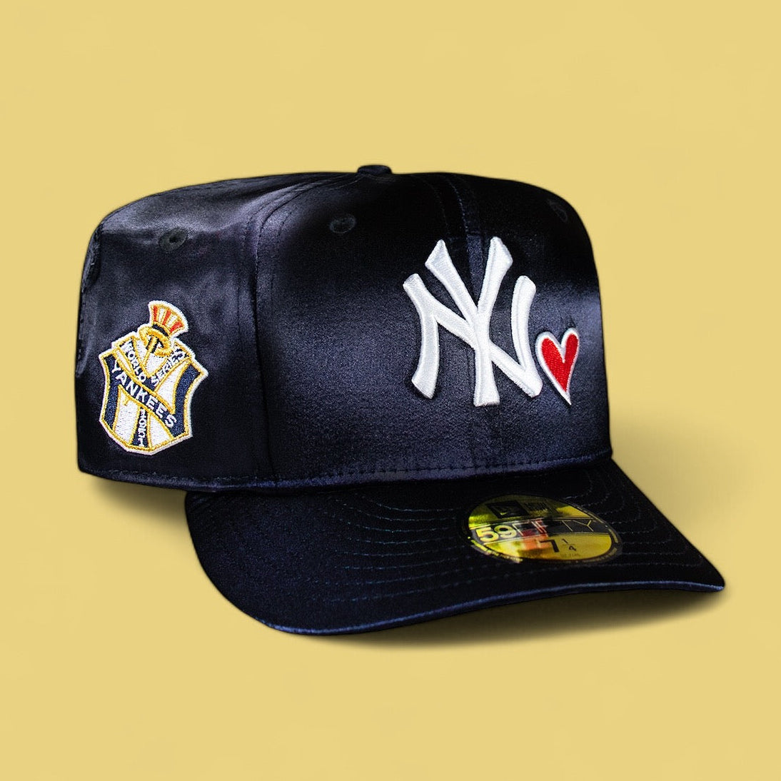 A navy satin baseball cap with the New York Yankees logo and a heart icon on the front, featuring a yellow and white badge on the side.